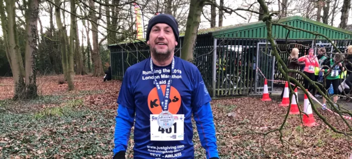 Andy is celebrating after the London Marathon. He is wearing a Kids t-shirt and his medal. He is in some kind of forest area.