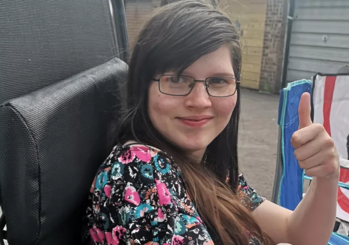 Erin is sitting on a lounge chair, giving a thumbs up to the camera. She is smiling and is wearing a dress with flower patterns.