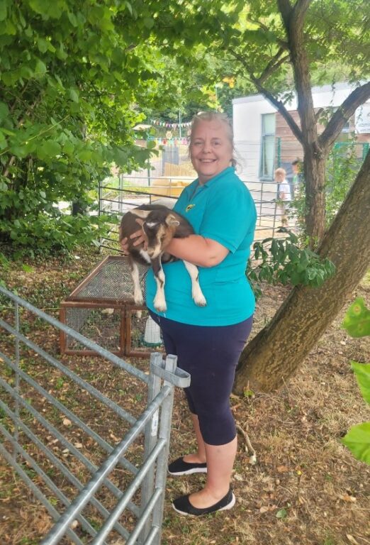 A staff member is standing under a tree and holding a goat in her arms. She is wearing a blue t-shirt and is looking at the camera, smiling.
