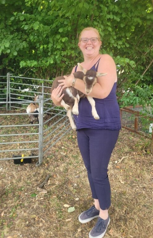 A woman is holding onto a baby goat. Behind them there are trees and an enclosure with another goat.