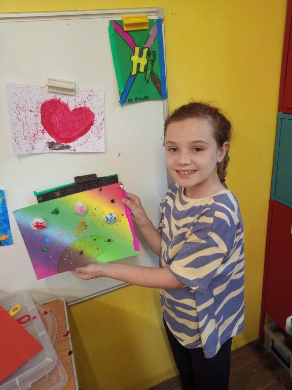 A child is holding up a painting they made. The painting has a rainbow background with fake butterflies layered on top, and splashes of paint.