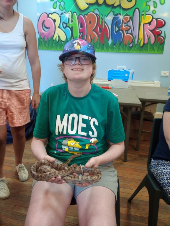 A child is sitting indoors on a chair. The child is holding a snake and smiling at the camera.