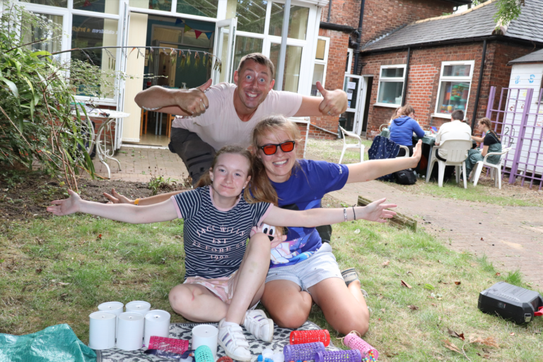 A young person and two staff members are sitting on the grass, cheering. In the background there are more young people sitting around a table.