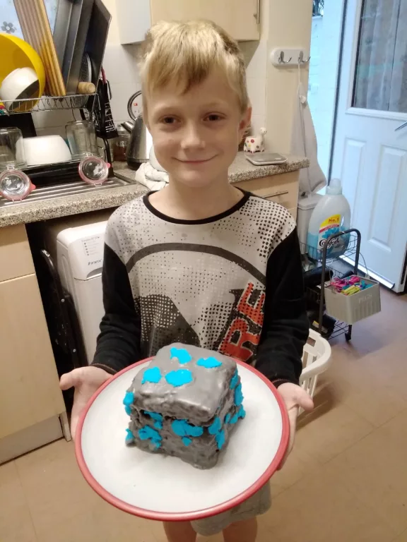 Chris is holding a cake covered in grey and blue icing. He is wearing a black and grey top.