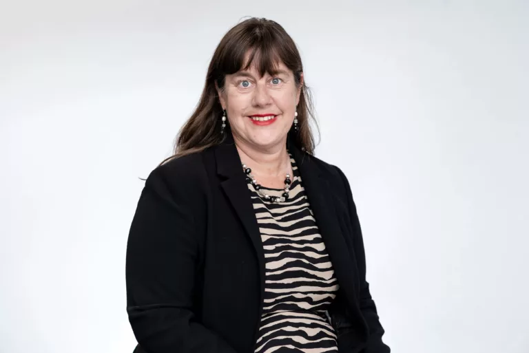 Diana Sutton is sitting for a professional photo. She has long brown hair and bangs. She is wearing a beige and black zebra top with a black blazer on top.