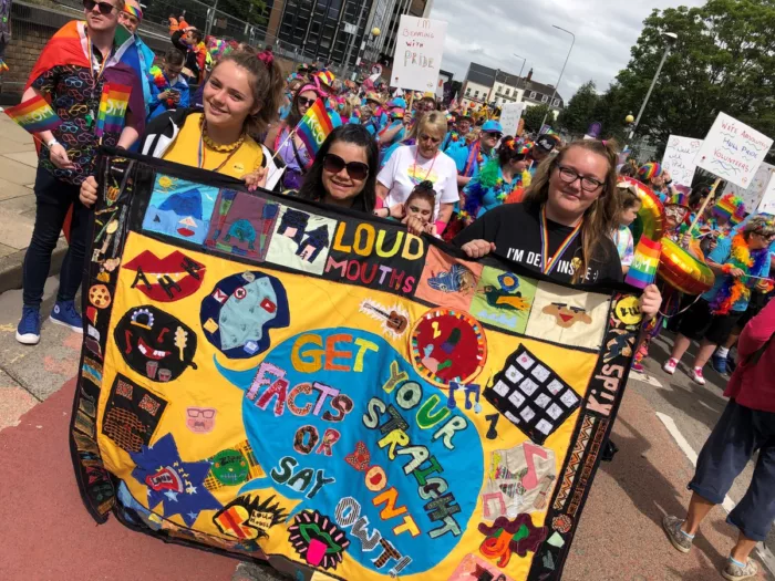 Three young people are at a Pride parade, holding a giant banner which reads 'Loud Mouths - get your facts straight or don't say owt'. The banner is decorated with colourful elements. In the background there are other people marching.