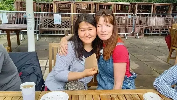 One woman is leaning on another woman, who has an arm wrapped around her. They are sitting at an outdoor table.