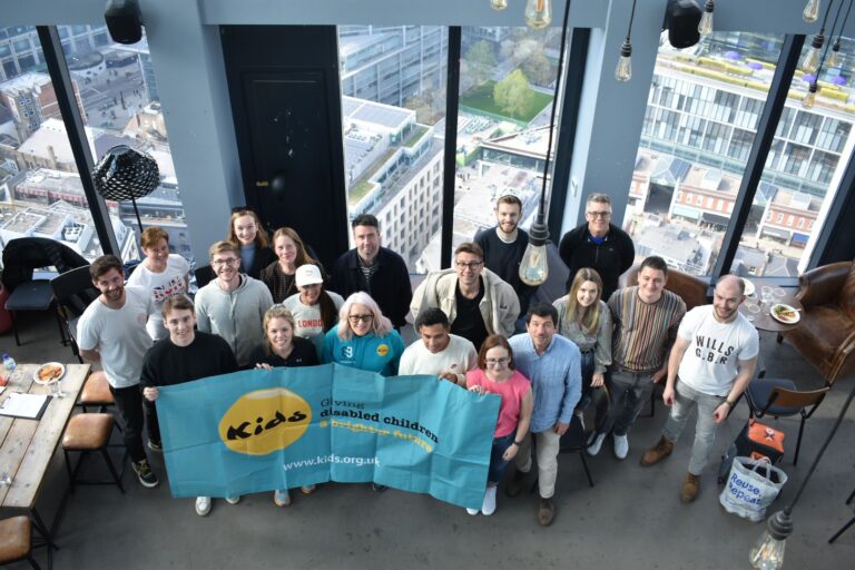 A large group of people are standing together by large windows. They are holding onto a blue Kids banner with the old logo on it.