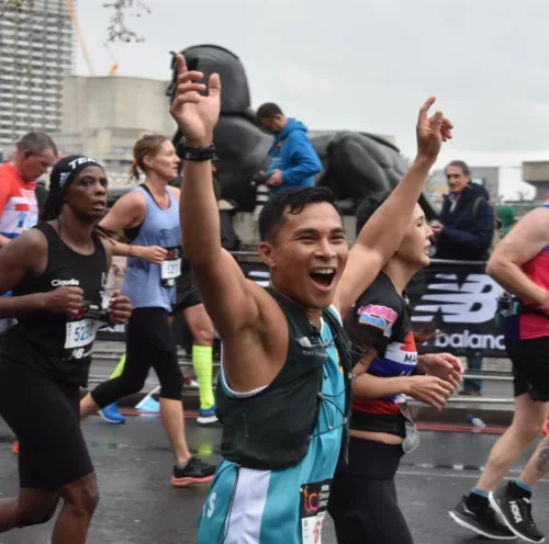A man is running the London Marathon. He is wearing a blue Kids vest and is raising both arms in the hair. He is smiling widely and is looking at someone off camera. In the background there are other people running.