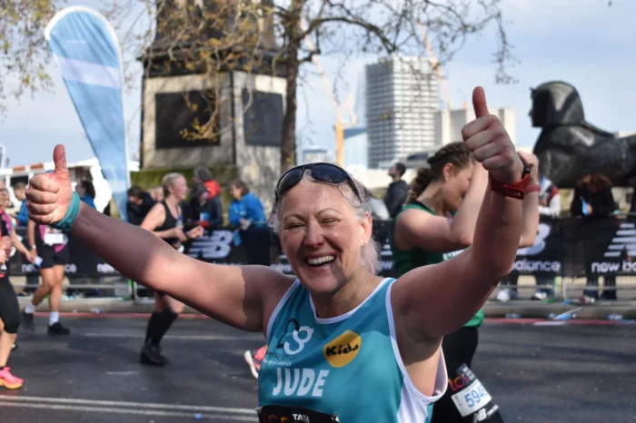 A London Marathon runner is running. She is holding her hands up in the air and is laughing. She is wearing a blue vest with the old Kids logo on it. In the background there are other people running.