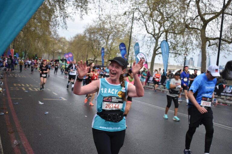 A London Marathon runner is waving at someone off camera. She seems to be saying something. She is wearing a blue vest with the Kids logo on it, and a black cap. In the background there are other runners.