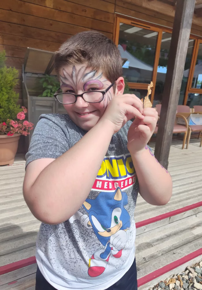 A child is standing outside, holding a brown leaf close to his face. He is wearing face paint and glasses.