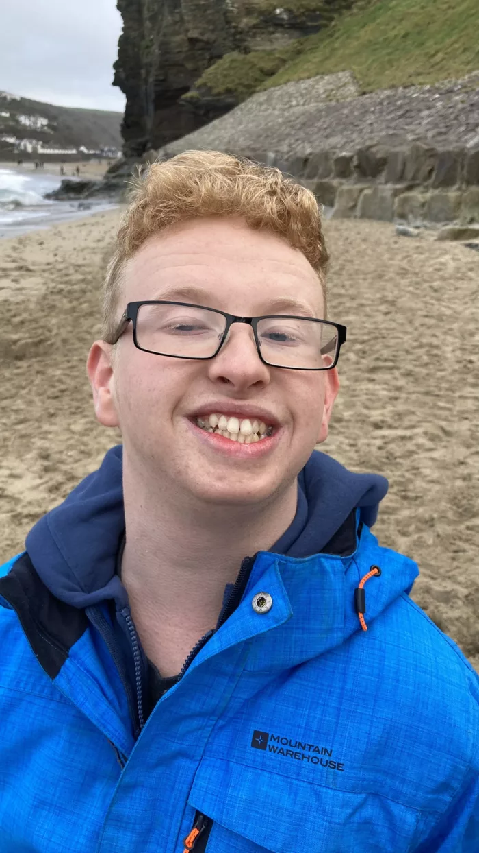 Nathan is at the beach, taking a selfie. He is wearing a bright blue coat and some eyeglasses.