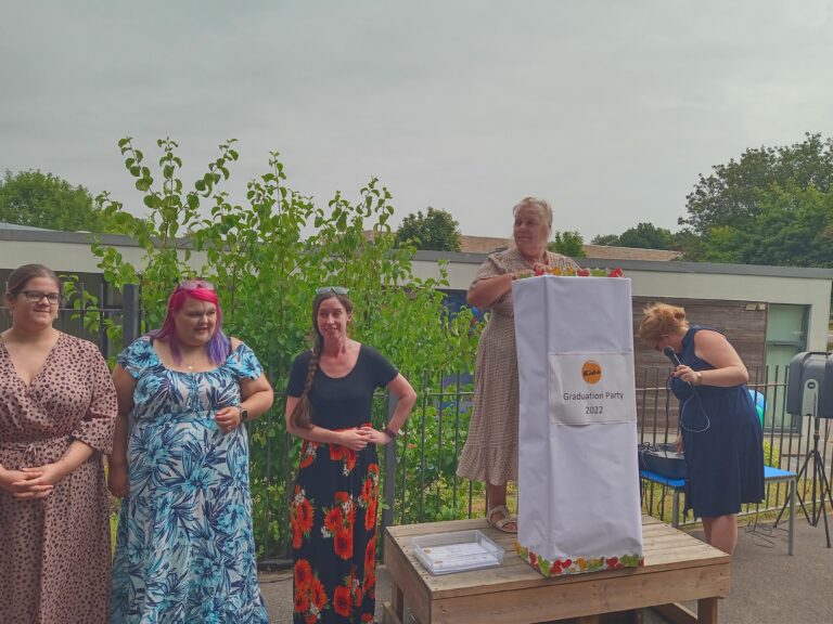 Staff members are hosting the graduation party. One staff member is standing on a wooden stage with a makeshift podium.