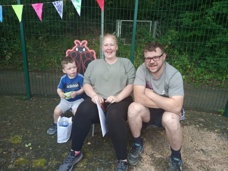 A family of three is sitting on a bench. Behind them there is a fence and a party flag banner.