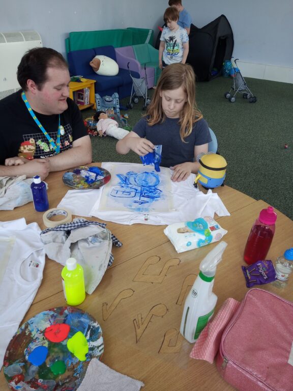 a child is sitting with a staff and making art. In the background there are two other children.