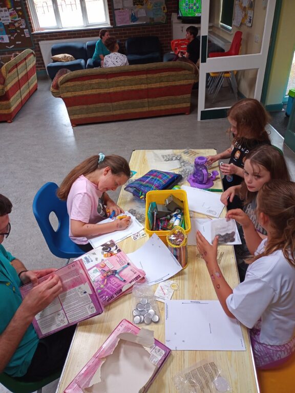 Young carers are sitting around a table, making badges. In the background there are young carers and a staff member sitting on sofas.