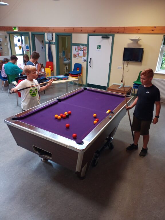 Two young carers are playing pool. In the background, more young carers are sitting around a table.