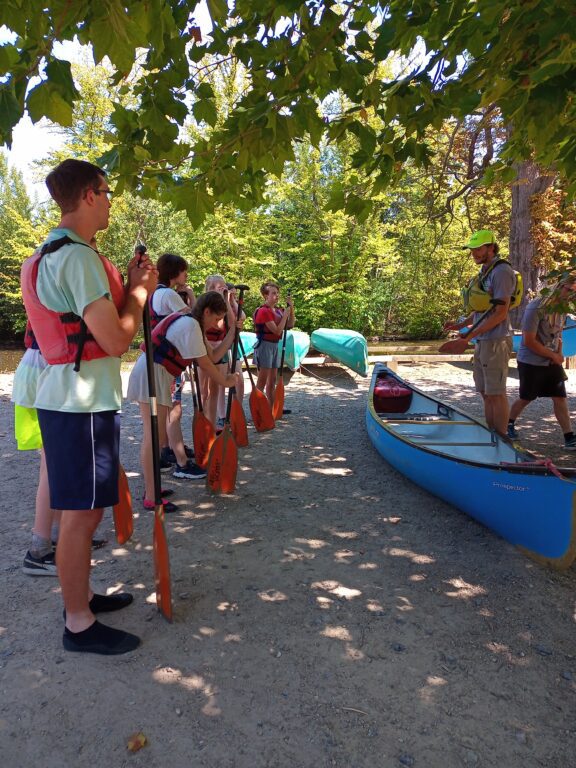 Young carers are standing in line while wearing lifejackets and holding onto paddles. In front of them there is a canoe and an instructor explaining how to use it.