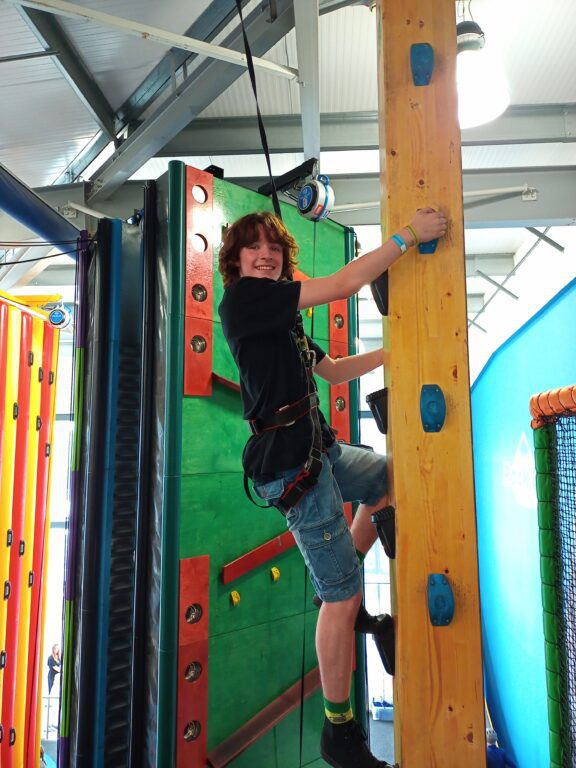 A young carer is taking on a climbing pole. The young carer is wearing knee-length shorts and a t-shirt. They are held up by a safety harness. 