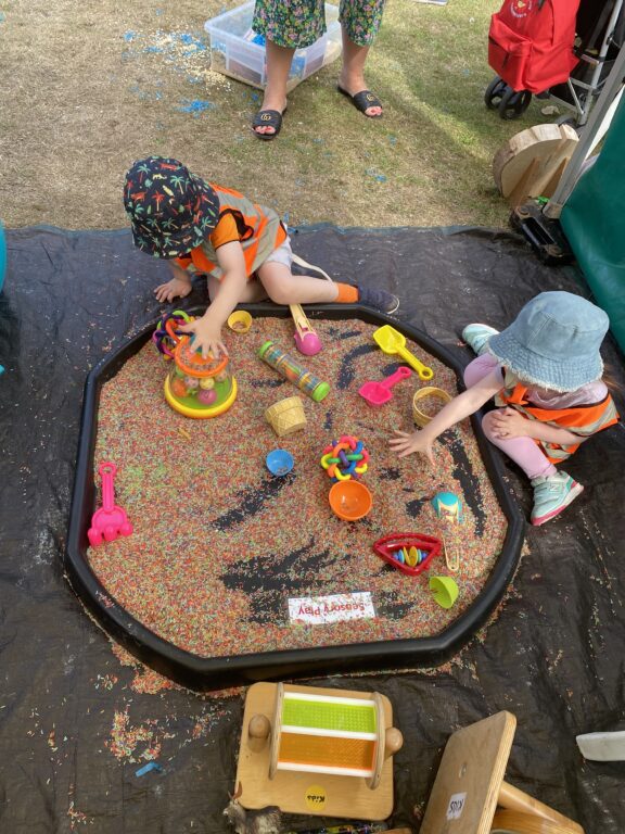 Two children are kneeling on the ground, playing with coloured rice and toys which sit in a large container.
