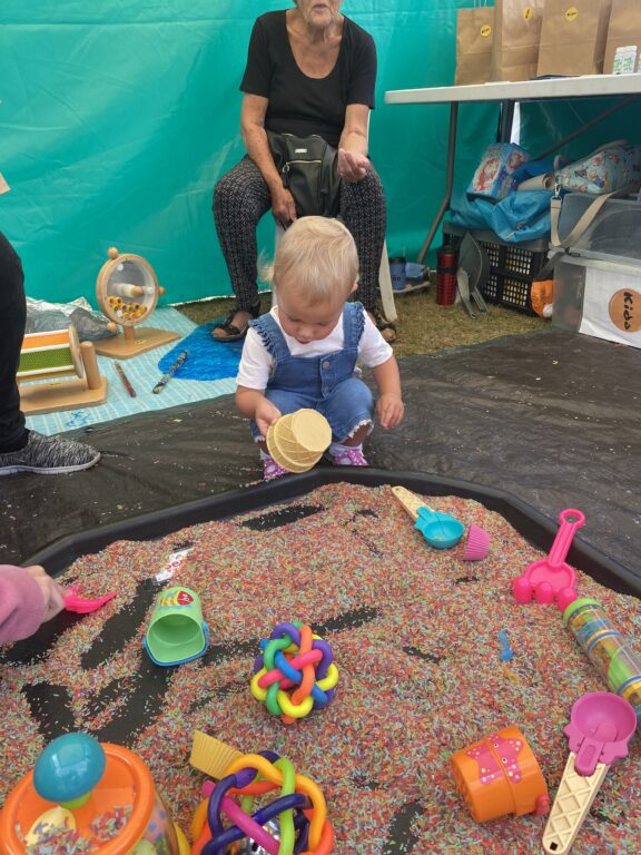 A child with blond hair is playing with coloured rice and toys which sit in a large floor container.