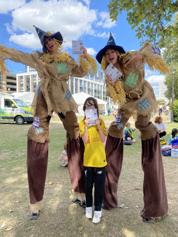 A child in a yellow t-shirt is standing between two women on stilts. They are dressed as scarecrows.