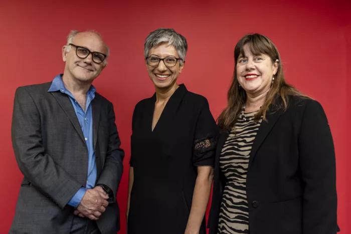 Previous Chair Stephen Unwin, CEO Katie Ghose, and current chair Diana Sutton are standing together in front of a red background. They are wearing smart clothes and are smiling.