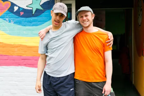 A young person and a staff member are standing with their arms over each other's shoulders. The young person is a wearing a grey t-shirt while the staff member is wearing an orange one. Both are wearing caps.