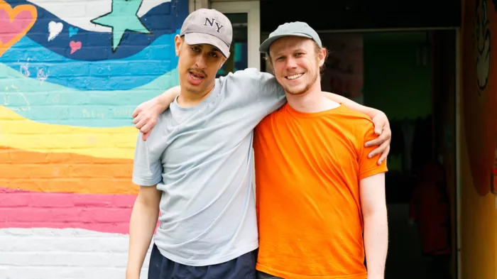 A young person and a staff member are standing with their arms over each other's shoulders. The young person is a wearing a grey t-shirt while the staff member is wearing an orange one. Both are wearing caps.