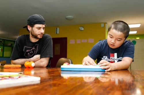 A boy is sitting around a table with a staff member. He is playing with a flat object that is obscured by the light. The staff member is looking at him.