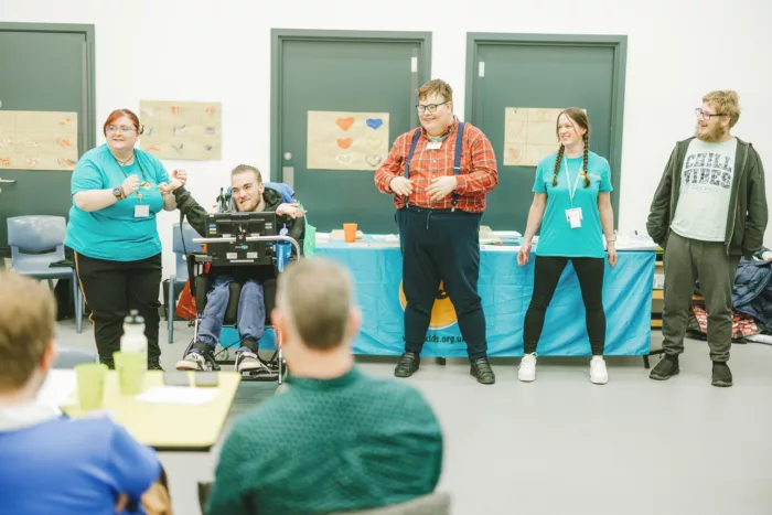 Five young people standing giving a presentation. Two are wearing Kids t-shirts, one is in a wheelchair.