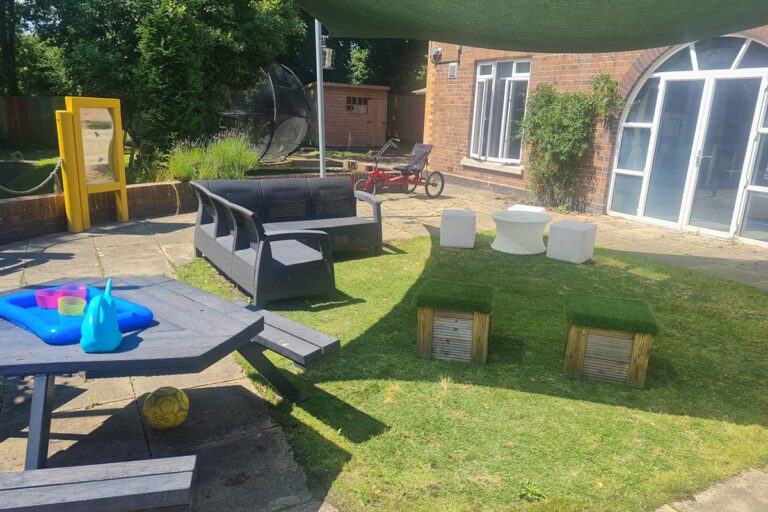 A bright sunny garden next to a brick house with big windows containing a picnic bench, seats, toys, a trike, a shed, and a sun canopy.