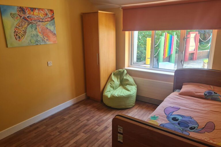 A bedroom painted orange with a double bed, bean bag, wardrobe. The bed has a duvet with cartoon characters and there is a bright painting of a turtle on the wall.