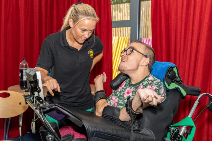 Jimmy is in his wheelchair, looking up at a staff member who is wearing a black polo t-shirt. There are red curtains behind them.