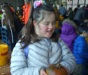 Taylor is at an event, holding a carved pumpkin in her hands. Her long brown hair in a ponytail and she is wearing a grey jacket and black trousers.
