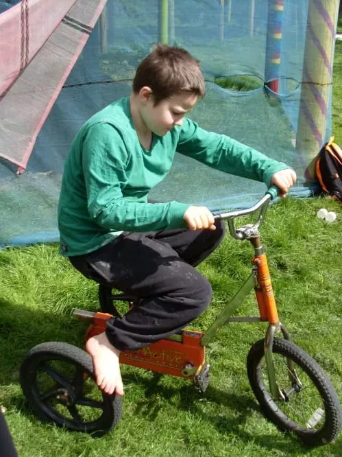Alex is riding a bicycle at the playground. He is wearing a green jumper and dark trousers.