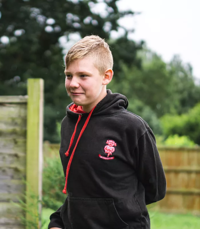 A photo of 14 year old Ben. He has tapered blonde hair and is wearing a black hoodie with red drawstrings and the Lincoln City F.C. logo on the right.