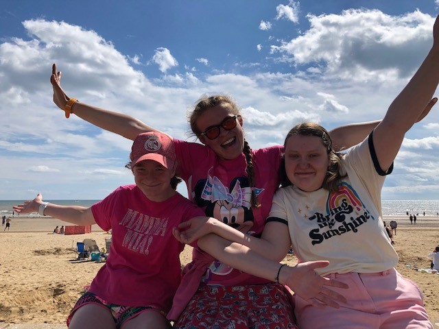 Three young people are sitting on a wall by the beach, with their arms up in the air. They are all smiling.