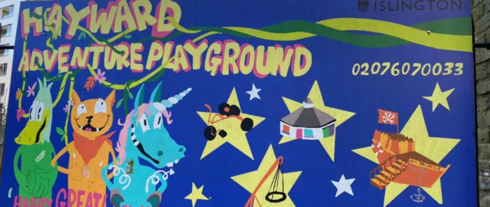A blue mural that reads, 'Hayward adventure playground'. On the mural there are animal characters and playground items including a bike and a playground castle.