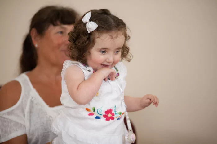 A child is walking around, one hand up to her chin. She is smiling and is wearing a white outfit with a matching bow in her hair. There is a woman behind her.