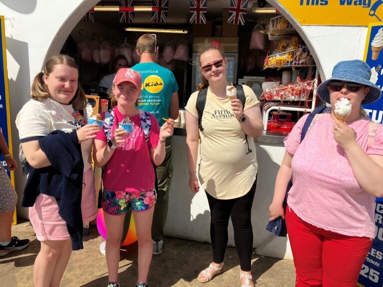 Four young people are standing in front of a shop, eating ice cream. There is a staff member behind them.