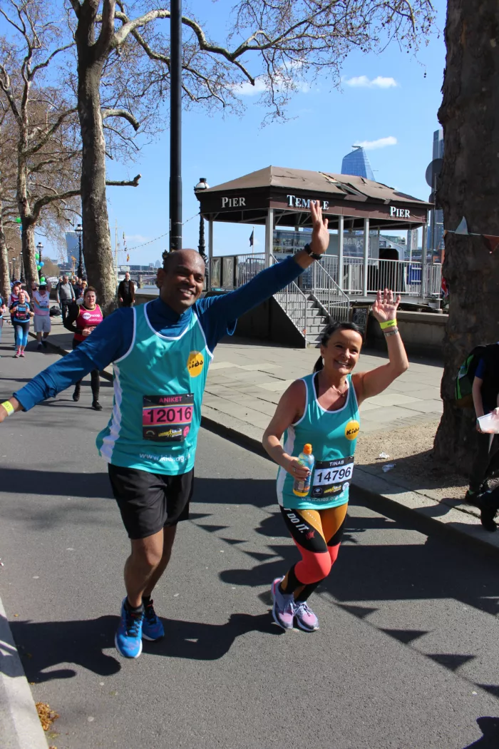 Two runners are taking part in the London Landmarks Half Marathon. They are wearing blue vests with the old Kids logo on them, and are waving at the camera.