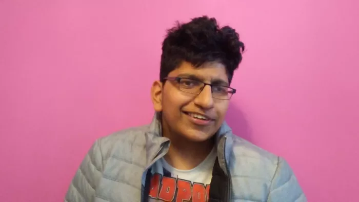 Karanjit is standing in front of a pink wall. He is wearing a grey coat and square eyeglasses. He has thick wavy hair.