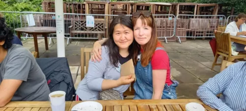 Two women are siting at a table, giving each other a side hug. They are smiling at the camera.