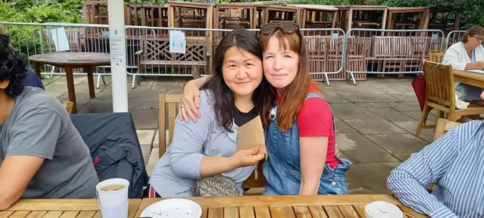 Two women are siting at a table, giving each other a side hug. They are smiling at the camera.