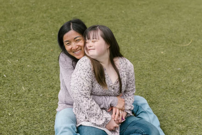An adult woman and teenage girl sit on grass hugging and smiling.