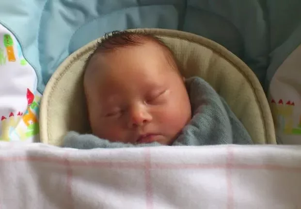 Baby Ollie is sleeping in his bed. There is a pink blanket over him.