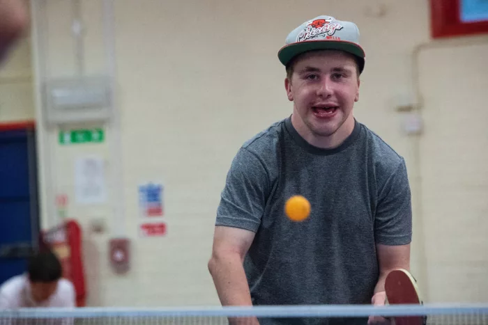 A young man is playing ping pong. He is about to hit a ball which is flying towards him. He is wearing a grey t-shirt and a cap.