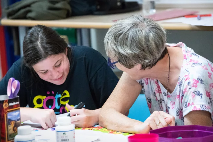 A young person is writing something down on paper. There is a staff member sitting next to the young person, smiling at them.